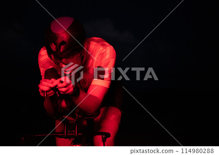 A triathlete rides his bike in the darkness of night, pushing himself to prepare for a marathon. The contrast between the darkness and the light of his bike creates a sense of drama and highlights the A triathlete rides his bike in the darkness of night, pushing himself to prepare for a marathon. The contrast between the darkness and the light of his bike creates a sense of drama and highlights the 114980288