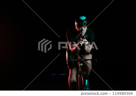 A triathlete rides his bike in the darkness of night, pushing himself to prepare for a marathon. The contrast between the darkness and the light of his bike creates a sense of drama and highlights the 114980304