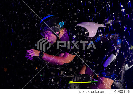 A triathlete braving the rain as he cycles through the night, preparing himself for the upcoming marathon. The blurred raindrops in the foreground and the dark, moody atmosphere in the background add 114980306