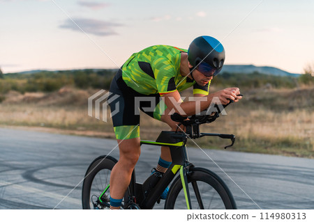 Triathlete riding his bicycle during sunset, preparing for a marathon. The warm colors of the sky provide a beautiful backdrop for his determined and focused effort. Triathlete riding his bicycle during sunset, preparing for a marathon. The warm colors of the sky provide a beautiful backdrop for his determined and focused effort. 114980313