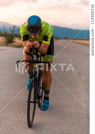 Triathlete riding his bicycle during sunset, preparing for a marathon. The warm colors of the sky provide a beautiful backdrop for his determined and focused effort. Triathlete riding his bicycle during sunset, preparing for a marathon. The warm colors of the sky provide a beautiful backdrop for his determined and focused effort. 114980316