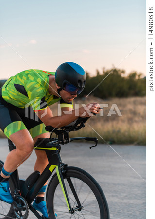 Triathlete riding his bicycle during sunset, preparing for a marathon. The warm colors of the sky provide a beautiful backdrop for his determined and focused effort. Triathlete riding his bicycle during sunset, preparing for a marathon. The warm colors of the sky provide a beautiful backdrop for his determined and focused effort. 114980318