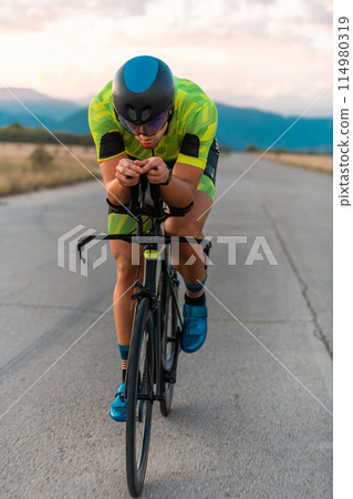 Triathlete riding his bicycle during sunset, preparing for a marathon. The warm colors of the sky provide a beautiful backdrop for his determined and focused effort. Triathlete riding his bicycle during sunset, preparing for a marathon. The warm colors of the sky provide a beautiful backdrop for his determined and focused effort. 114980319