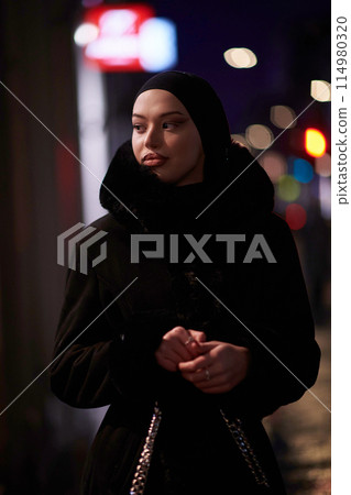 Muslim woman walking on an urban city street on a cold winter night wearing hijab Muslim woman walking on an urban city street on a cold winter night wearing hijab 114980320