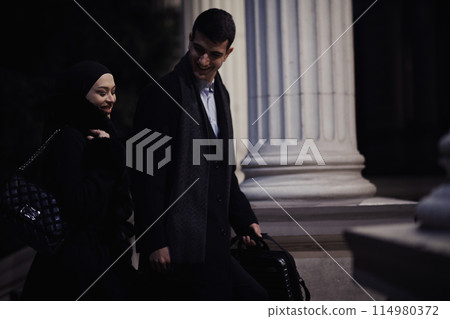 Happy multicultural business couple walking together outdoors in an urban city street at night near a jewelry shopping store window. Happy multicultural business couple walking together outdoors in an urban city street at night near a jewelry shopping store window. 114980372