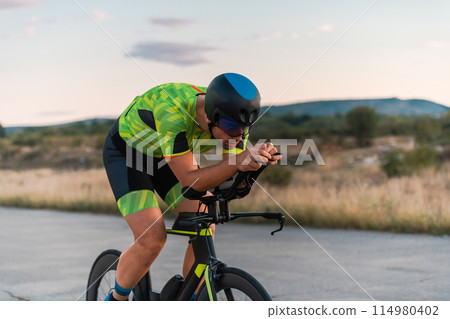 Triathlete riding his bicycle during sunset, preparing for a marathon. The warm colors of the sky provide a beautiful backdrop for his determined and focused effort. 114980402