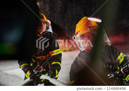 Firefighters using a water hose to eliminate a fire hazard. Team of female and male firemen in dangerous rescue mission.  114980406