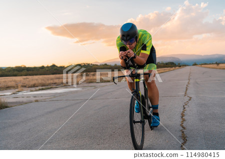 Triathlete riding his bicycle during sunset, preparing for a marathon. The warm colors of the sky provide a beautiful backdrop for his determined and focused effort. 114980415