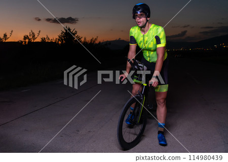 A triathlete rides his bike in the darkness of night, pushing himself to prepare for a marathon. The contrast between the darkness and the light of his bike creates a sense of drama and highlights the 114980439