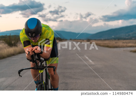 Triathlete riding his bicycle during sunset, preparing for a marathon. The warm colors of the sky provide a beautiful backdrop for his determined and focused effort. Triathlete riding his bicycle during sunset, preparing for a marathon. The warm colors of the sky provide a beautiful backdrop for his determined and focused effort. 114980474