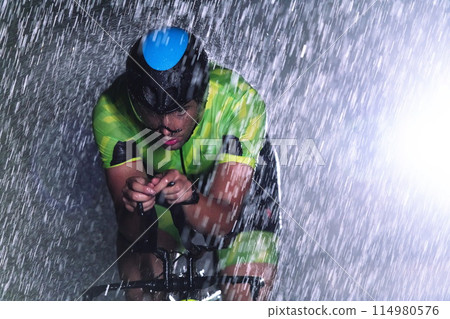 A triathlete braving the rain as he cycles through the night, preparing himself for the upcoming marathon. The blurred raindrops in the foreground and the dark, moody atmosphere in the background add 114980576