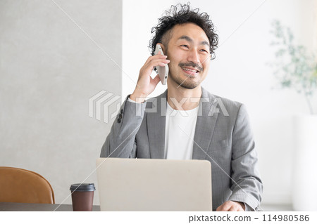 A man talking on a smartphone in an office while using a computer A man talking on a smartphone in an office while using a computer 114980586