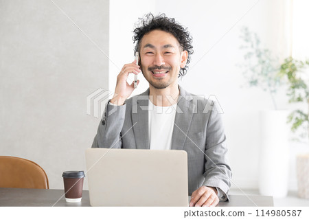 A man talking on a smartphone in an office while using a computer A man talking on a smartphone in an office while using a computer 114980587