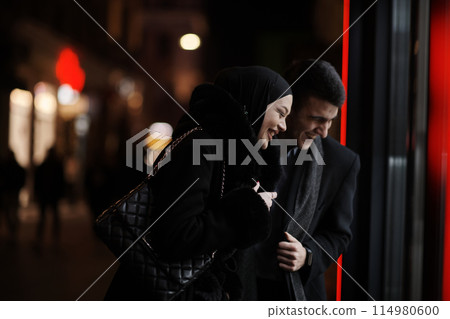 Happy multicultural business couple walking together outdoors in an urban city street at night near a jewelry shopping store window. Happy multicultural business couple walking together outdoors in an urban city street at night near a jewelry shopping store window. 114980600
