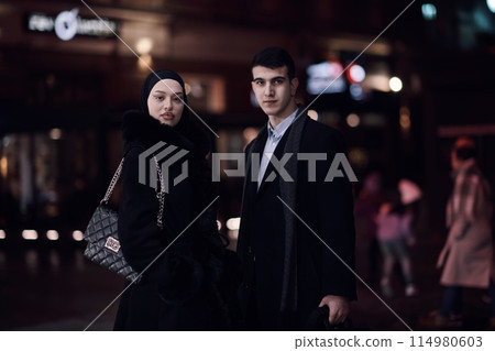 Happy multicultural business couple walking together outdoors in an urban city street at night near a jewelry shopping store window. Happy multicultural business couple walking together outdoors in an urban city street at night near a jewelry shopping store window. 114980603