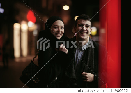 Happy multicultural business couple walking together outdoors in an urban city street at night near a jewelry shopping store window. Happy multicultural business couple walking together outdoors in an urban city street at night near a jewelry shopping store window. 114980622