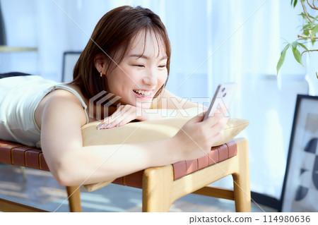 A woman lying on a chair in a room and looking at her smartphone 114980636