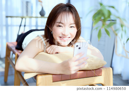 A woman lying on a chair in a room and looking at her smartphone A woman lying on a chair in a room and looking at her smartphone 114980650