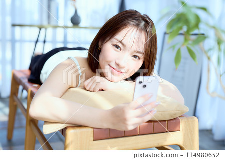 A woman lying on a chair in a room and looking at her smartphone 114980652