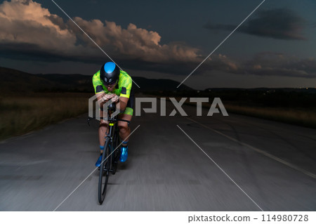 A triathlete rides his bike in the darkness of night, pushing himself to prepare for a marathon. The contrast between the darkness and the light of his bike creates a sense of drama and highlights the A triathlete rides his bike in the darkness of night, pushing himself to prepare for a marathon. The contrast between the darkness and the light of his bike creates a sense of drama and highlights the 114980728