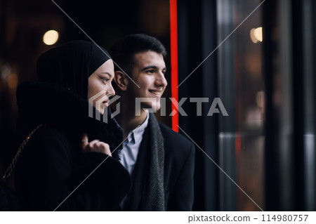 Happy multicultural business couple walking together outdoors in an urban city street at night near a jewelry shopping store window. Happy multicultural business couple walking together outdoors in an urban city street at night near a jewelry shopping store window. 114980757