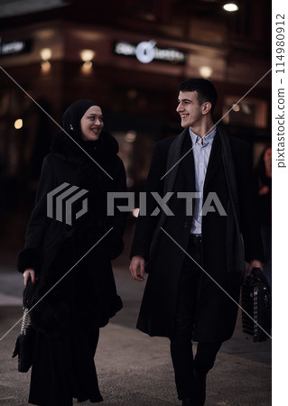 Happy multicultural business couple walking together outdoors in an urban city street at night near a jewelry shopping store window. Happy multicultural business couple walking together outdoors in an urban city street at night near a jewelry shopping store window. 114980912