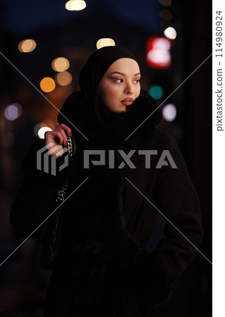 Muslim woman walking on an urban city street on a cold winter night wearing hijab Muslim woman walking on an urban city street on a cold winter night wearing hijab 114980924