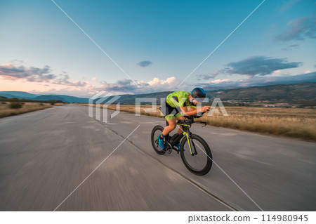 Triathlete riding his bicycle during sunset, preparing for a marathon. The warm colors of the sky provide a beautiful backdrop for his determined and focused effort. Triathlete riding his bicycle during sunset, preparing for a marathon. The warm colors of the sky provide a beautiful backdrop for his determined and focused effort. 114980945