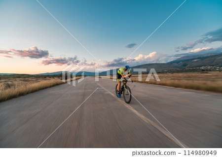 Triathlete riding his bicycle during sunset, preparing for a marathon. The warm colors of the sky provide a beautiful backdrop for his determined and focused effort. 114980949