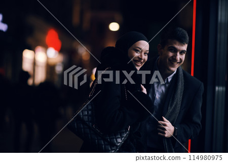 Happy multicultural business couple walking together outdoors in an urban city street at night near a jewelry shopping store window. Happy multicultural business couple walking together outdoors in an urban city street at night near a jewelry shopping store window. 114980975