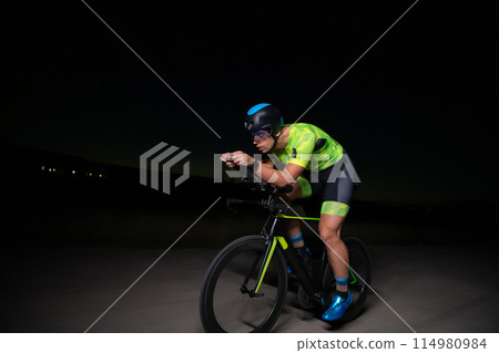 A triathlete rides his bike in the darkness of night, pushing himself to prepare for a marathon. The contrast between the darkness and the light of his bike creates a sense of drama and highlights the A triathlete rides his bike in the darkness of night, pushing himself to prepare for a marathon. The contrast between the darkness and the light of his bike creates a sense of drama and highlights the 114980984
