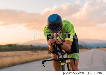 Triathlete riding his bicycle during sunset, preparing for a marathon. The warm colors of the sky provide a beautiful backdrop for his determined and focused effort. Triathlete riding his bicycle during sunset, preparing for a marathon. The warm colors of the sky provide a beautiful backdrop for his determined and focused effort. 114980986