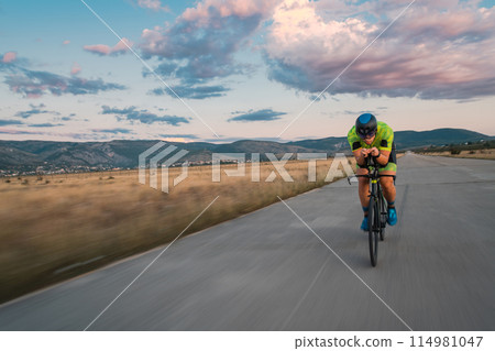 Triathlete riding his bicycle during sunset, preparing for a marathon. The warm colors of the sky provide a beautiful backdrop for his determined and focused effort. 114981047