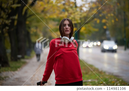 Portrait of running woman after jogging in the park on autumn seasson. Female fitness model training outside on a cozy fall day and listening to music over smartphone. 114981106
