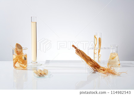Front view photograph on white background, an empty podium placed in center with a ginseng leaning on, beside a measuring cylinder, beaker, test tube and erlenmeyer, all items containing yellow fluid 114982543