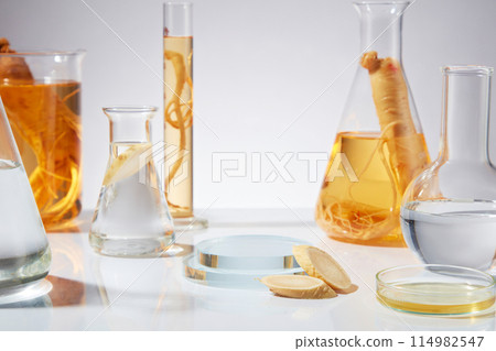 Close-up front view photo of ginseng ingredient theme in laboratory, ginseng extract and transparent liquid contained in erlenmeyer and boiling flask, glass podium in center for product presentation 114982547