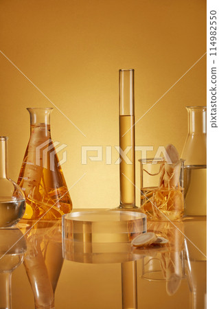 Yellow color tone photo with ginseng experiment theme, glassware in lab containing yellow liquid and ginseng root decorated behind a round glass pedestal. Frontal shot photo with space for text Yellow color tone photo with ginseng experiment theme, glassware in lab containing yellow liquid and ginseng root decorated behind a round glass pedestal. Frontal shot photo with space for text 114982550