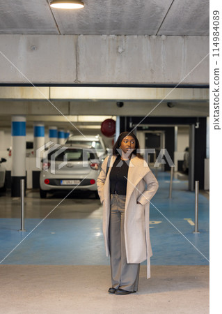 Stylish woman in elegant coat poses confidently in a parking garage, embodying casual urban fashion 114984089