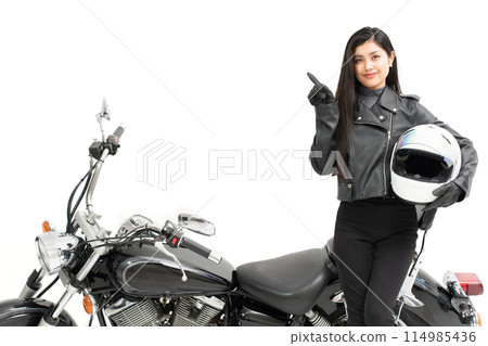 A biker girl standing next to her bike, holding a helmet and pointing 114985436