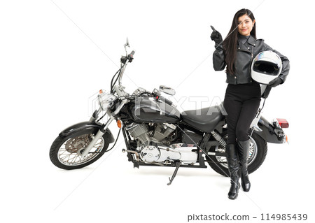 A biker girl standing next to her bike, holding a helmet and pointing 114985439
