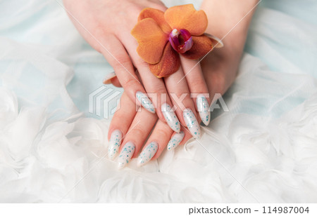 Showcasing Floral Nail Art and Holding an Orange Blossom on a Soft Textured Surface 114987004