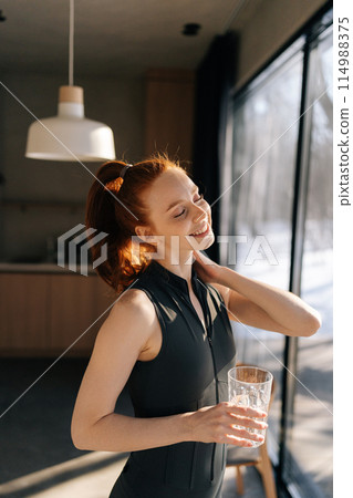 Vertical portrait of athletic young woman in sport outfit standing by window and drinking fresh water in morning after fitness training workout exercise at home, enjoying sunny weather and lifestyle. 114988375