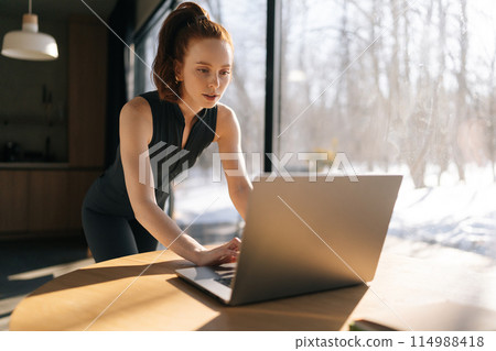 Focused sporty young woman in outfit using laptop standing at desk by window on sunny day, thinking of online creative task, browsing internet, search information, working on computer typing email Focused sporty young woman in outfit using laptop standing at desk by window on sunny day, thinking of online creative task, browsing internet, search information, working on computer typing email 114988418