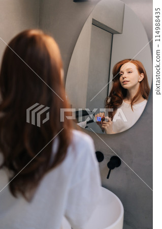 Vertical shot of young woman playing with long red hair looking in mirror standing in bathroom in morning. Reflection on mirror of pretty redhead female preens, admires, straightening hairstyle Vertical shot of young woman playing with long red hair looking in mirror standing in bathroom in morning. Reflection on mirror of pretty redhead female preens, admires, straightening hairstyle 114988435