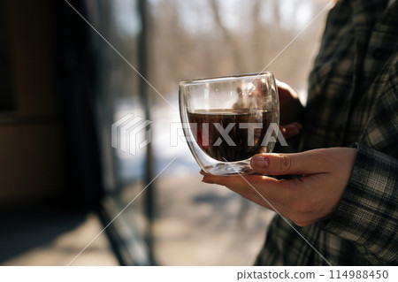 Closeup cropped shot of unrecognizable woman holding cup with hot aroma coffee in morning sunlight. Closeup of lady enjoying drinking coffee at home. Happy relaxed female drinking tea on kitchen. Closeup cropped shot of unrecognizable woman holding cup with hot aroma coffee in morning sunlight. Closeup of lady enjoying drinking coffee at home. Happy relaxed female drinking tea on kitchen. 114988450