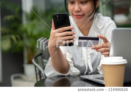 A young Asian woman holding a credit card and her smartphone while sitting at a table outdoors. A young Asian woman holding a credit card and her smartphone while sitting at a table outdoors. 114990826