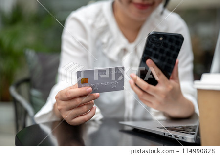 A young Asian woman holding a credit card and her smartphone while sitting at a table outdoors. 114990828