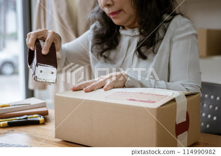 A mature Asian female online seller packing a shipping cardboard box, using a packing tape dispenser 114990982
