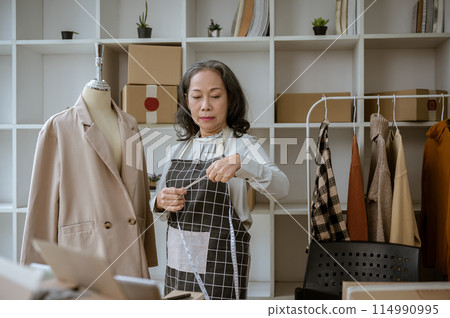 A mature Asian female tailor measuring a suit on a mannequin with a measuring tape. A mature Asian female tailor measuring a suit on a mannequin with a measuring tape. 114990995