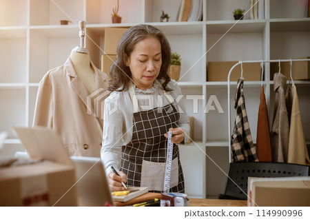 A mature Asian female tailor is taking notes on a notepad, measuring and adjusting the pattern. 114990996